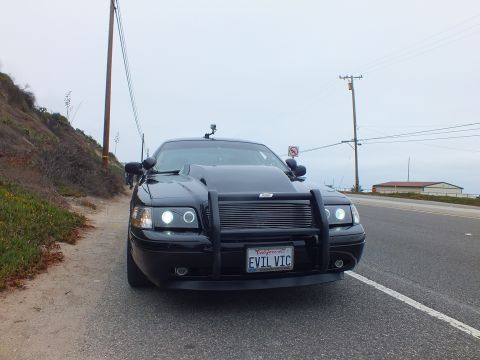2003 Ford Crown Victoria - Photograph of a blacked-out Crown Victoria with a push bar and 'EVIL VIC' license plate parked on a coastal road, not a technical document.