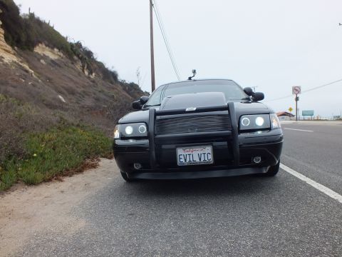 2003 Ford Crown Victoria - Low-angle photograph of a black Crown Victoria with an 'EVIL VIC' plate parked on a coastal road, not a technical document.