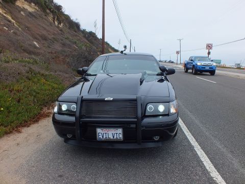 2003 Ford Crown Victoria - Photograph of a black Crown Victoria with a push bar and 'EVIL VIC' license plate on a coastal road, not a technical document.
