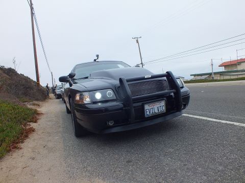 2003 Ford Crown Victoria - Photograph of a black Crown Victoria with a push bar and 'EVIL VIC' plate parked roadside, not a technical document.