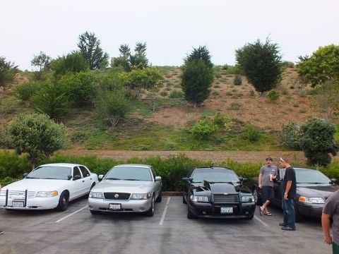 2003 Ford Crown Victoria - Photograph of four sedans parked in a lot below a hillside with two men standing nearby, not a technical document.