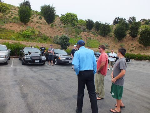 2003 Ford Crown Victoria - Photo of attendees standing and talking at a car meet in a lot with sedans parked along a landscaped hillside.