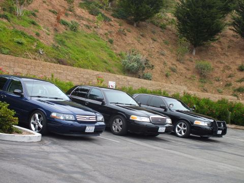 2003 Ford Crown Victoria - Photo of three full-size sedans parked at an angle in a parking lot below a hillside.