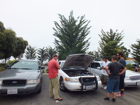 2003 Ford Crown Victoria - Photograph of people inspecting a sedan engine bay at a car meetup, not a technical document.