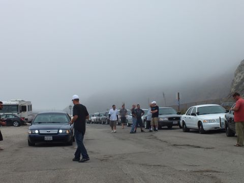 2003 Ford Crown Victoria - People mingle beside a row of parked cars, including white and dark sedans and an RV, at a fog-covered gravel lot.
