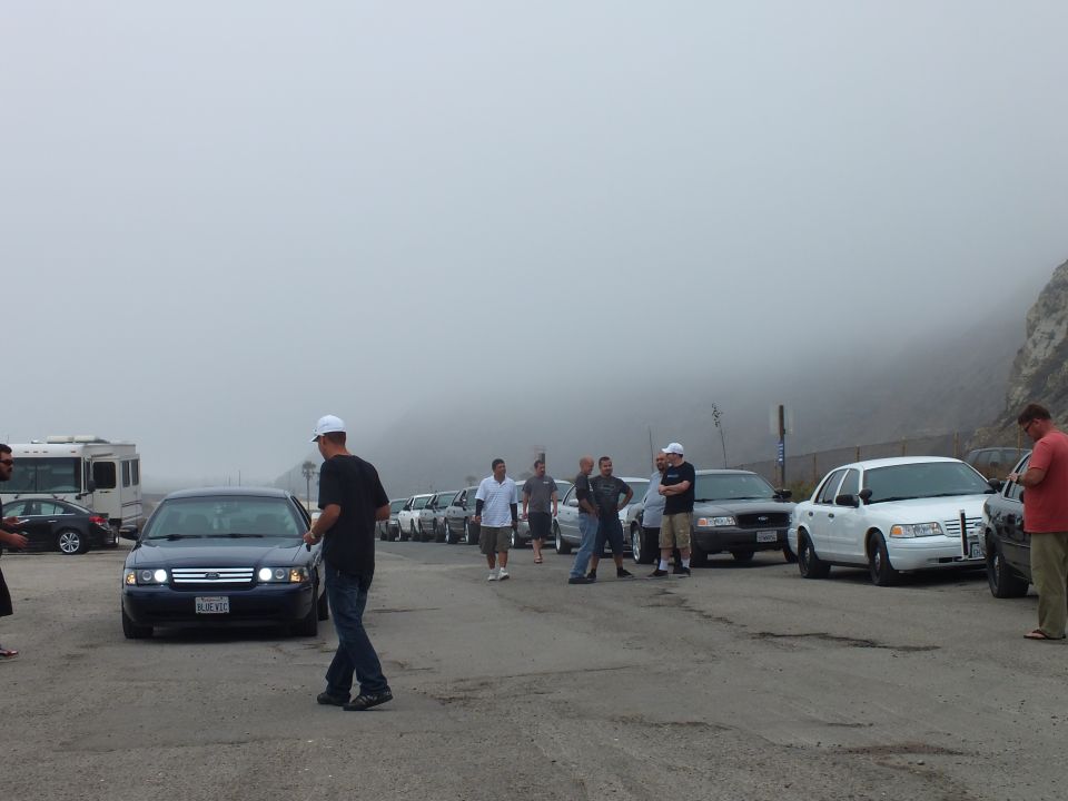 People and a row of parked cars including white and dark sedans and an RV at a foggy lot