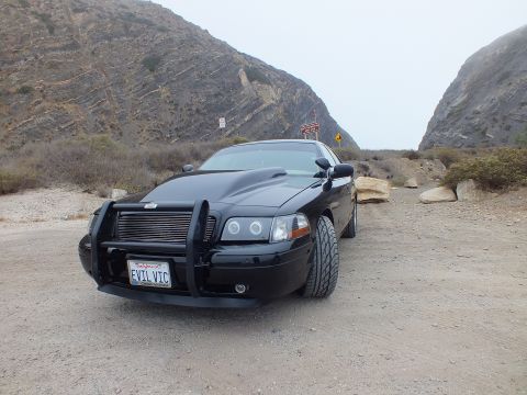 2003 Ford Crown Victoria - black customized sedan with a grille guard and 'EVIL VIC' plate poses on gravel within a fog-touched rocky canyon.