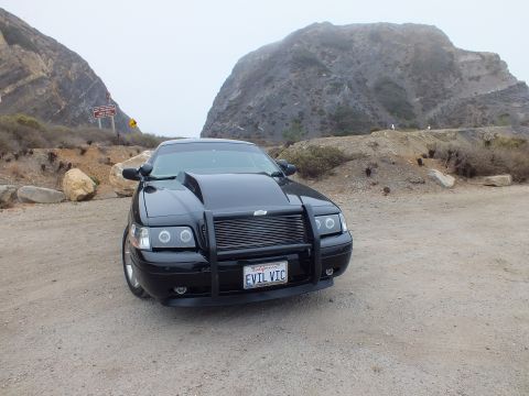 2003 Ford Crown Victoria - black customized sedan with a grille guard and 'EVIL VIC' plate faces the camera on gravel before a large rock outcrop.