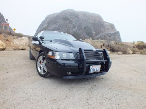 2003 Ford Crown Victoria - black customized sedan with a grille guard and red brake calipers sits on gravel near a large coastal rock outcrop.