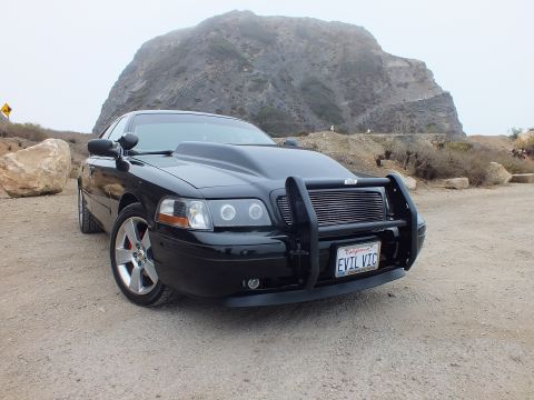 2003 Ford Crown Victoria - black Ford sedan with a grille guard and 'EVIL VIC' plate poses on gravel in front of a large fog-capped rock outcrop.