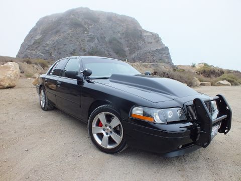 2003 Ford Crown Victoria - black Ford Crown Victoria sedan fitted with a push-bar brush guard sits on gravel below a fog-shrouded rocky headland.