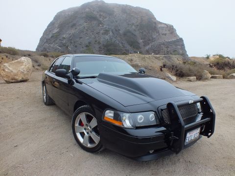 2003 Ford Crown Victoria - black Ford Crown Victoria with a chrome-grille push bar and California license plate parked on dirt under a misty coastal bluff.