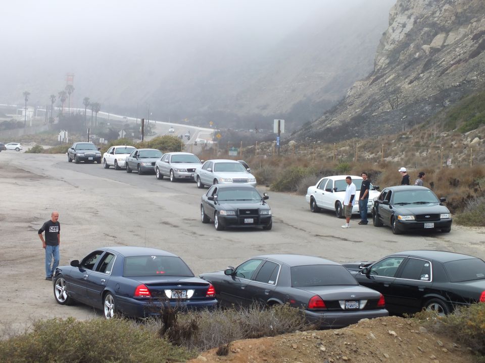 Sedans and a pickup parked at a foggy coastal lot with people standing nearby