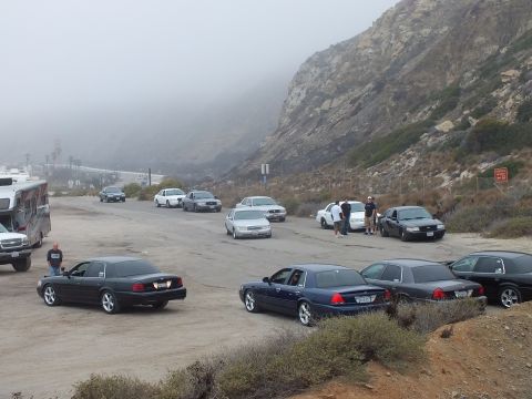 2003 Ford Crown Victoria - Photograph of a car meetup with sedans, an RV, and attendees at a foggy coastal parking lot; not a technical document.