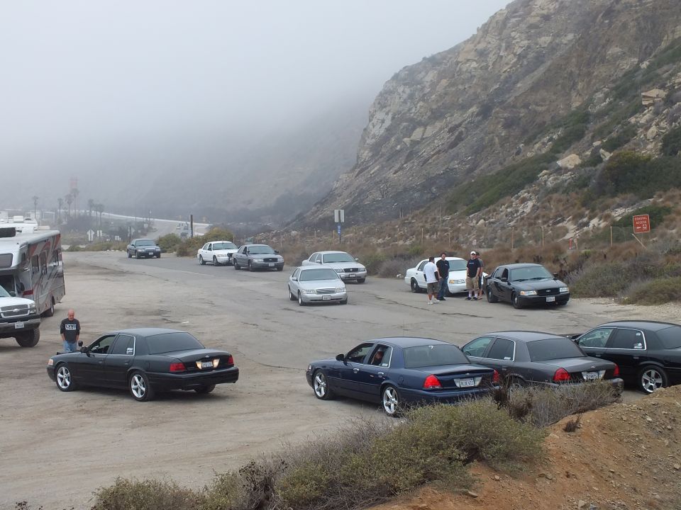Sedans parked at a foggy coastal lot with people and an RV