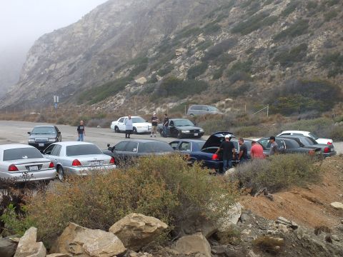 2003 Ford Crown Victoria - Full-size sedans parked in a foggy canyon pullout, one with its trunk open, as people gather among them.