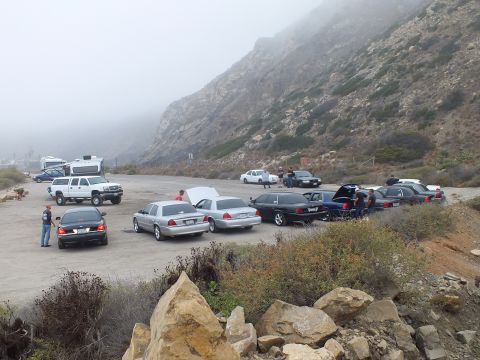 2003 Ford Crown Victoria - Sedans and pickups arranged across a gravel canyon lot with people standing beside open trunks under fog.