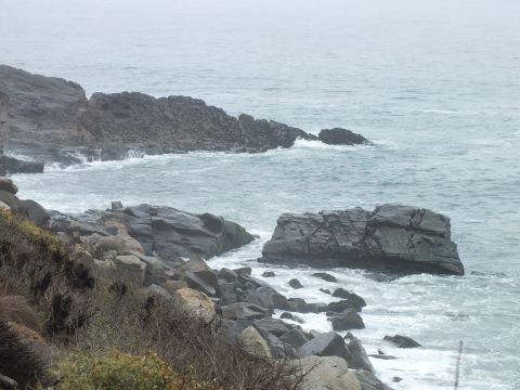 2003 Ford Crown Victoria - Surf breaks among gray boulders along a rugged rocky shoreline beneath an overcast sky.