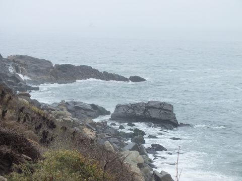 2003 Ford Crown Victoria - Rocky outcrops jut into the gray sea as waves break beneath a foggy pale sky along the coast.