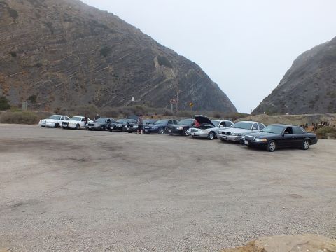 2003 Ford Crown Victoria - long row of full-size sedans parked across a wide dirt lot framed by two foggy mountain slopes.