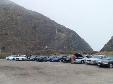 2003 Ford Crown Victoria - line of dark and white full-size sedans parked at the base of a steep foggy hillside, one hood open.