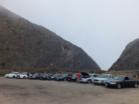 2003 Ford Crown Victoria - Full-size sedans lined up across a dirt lot beneath a steep foggy mountain, a person checking under a hood.