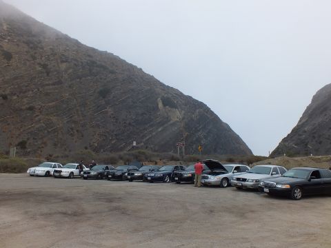 2003 Ford Crown Victoria - lineup of full-size sedans in a gravel lot at the foot of foggy mountain slopes, hood raised on one car.