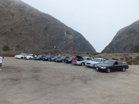 2003 Ford Crown Victoria - Sedans and a white pickup lined up across a dirt lot below foggy peaks as people stand nearby.