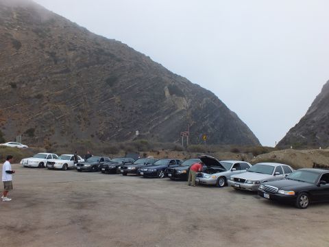 2003 Ford Crown Victoria - row of full-size sedans parked in a dirt lot at the base of a foggy ridge, one hood open and a person looking in.
