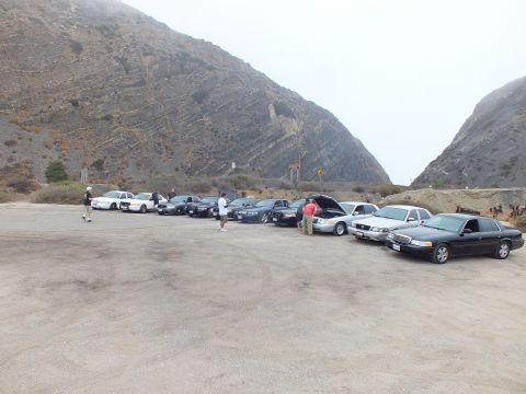2003 Ford Crown Victoria - line of sedans and a white pickup parked in a dirt lot at the base of foggy coastal hills, with several people standing among the cars.