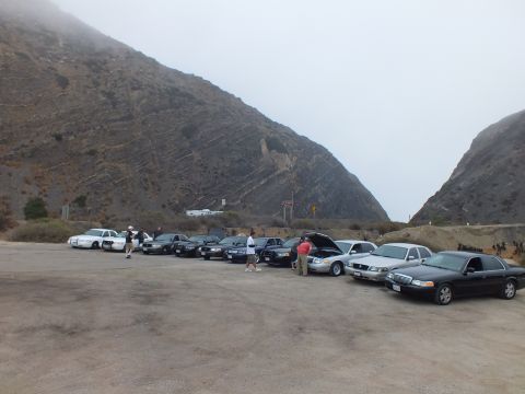 2003 Ford Crown Victoria - row of sedans and a white pickup parked in a foggy dirt lot below steep hills, with one car's hood raised and people standing nearby.