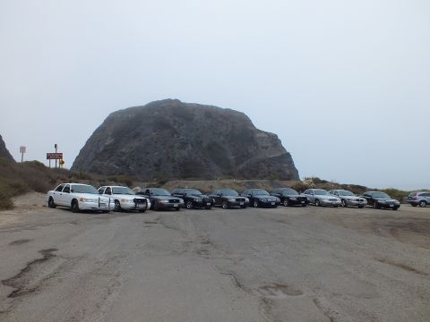 2003 Ford Crown Victoria - row of dark and white sedans plus a white pickup parked on cracked pavement facing a massive rounded rock outcrop under gray sky.