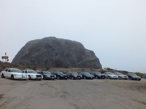 2003 Ford Crown Victoria - diagonal row of sedans, including white and black models, parked on worn pavement in front of a large rock formation on a foggy day.