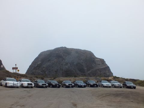 2003 Ford Crown Victoria - row of light and dark sedans parked on worn pavement in front of a large grayish rock formation under a flat gray sky.