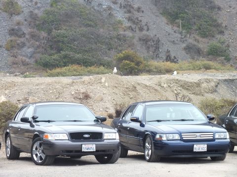 2003 Ford Crown Victoria - Two dark Ford sedans parked side by side in a dirt lot, the right one wearing a California plate reading BLUE VIC, with a gull on the bank…