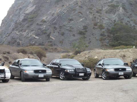 2003 Ford Crown Victoria - Three Ford sedans, one black with a push bumper, parked on gravel at the base of a steep rocky hillside.