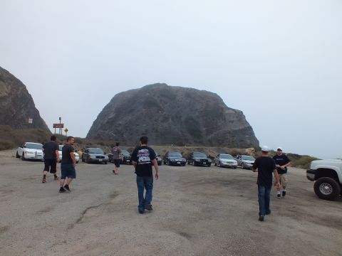 2003 Ford Crown Victoria - Several people in dark shirts walk across a paved lot toward a row of parked sedans at the base of a large rock formation on a hazy day.