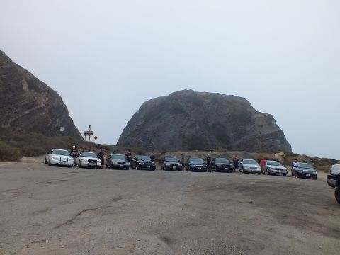 2003 Ford Crown Victoria - group of people stand among a row of light and dark sedans parked in a dirt lot at the base of a large rock formation under gray sky.