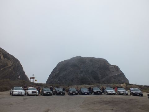 2003 Ford Crown Victoria - group of people stand among a wide row of parked sedans in a dirt lot below a large rounded rock formation under an overcast sky.