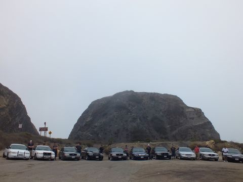 2003 Ford Crown Victoria - People pose alongside a long row of parked sedans in a dirt lot at the base of a large rock formation beneath a flat gray sky.