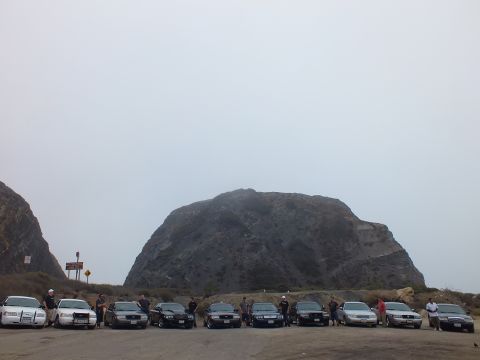 2003 Ford Crown Victoria - group of people stand among a row of light and dark sedans parked in a dirt lot below a large rock formation on a hazy day.