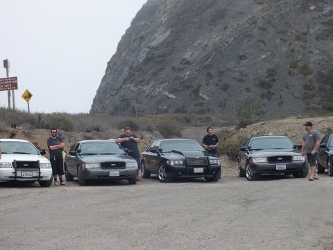 2003 Ford Crown Victoria - Several men stand beside four dark Ford sedans, one with a push bumper, parked in a dirt lot near a recreation info sign below a steep hill.