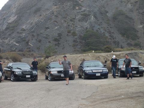 2003 Ford Crown Victoria - Group of people gathered beside several black full-size sedans parked in a gravel lot at the foot of a steep rocky hillside.