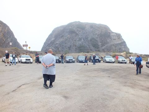2003 Ford Crown Victoria - People stand in a large gravel lot beside a row of parked sedans, with a massive fog-shrouded rock formation rising behind them.