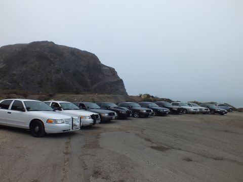 2003 Ford Crown Victoria - white full-size sedan with a front push bar leads an angled row of parked sedans beside a rocky hillside.