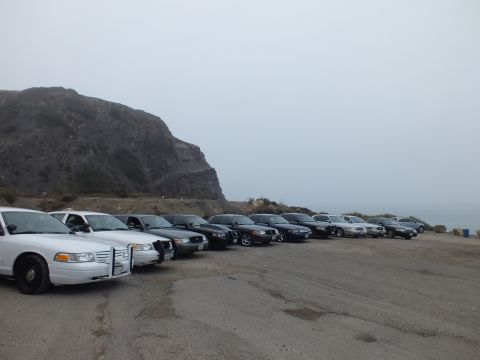 2003 Ford Crown Victoria - Full-size sedans parked at an angle on gravel beside a rocky coastal hill under overcast, foggy sky.