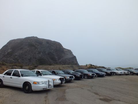 2003 Ford Crown Victoria - white full-size sedan with a push bar fronts a parked row of cars in a gravel lot below a large rocky hill.