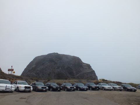 2003 Ford Crown Victoria - Full-size sedans parked in a row beneath a dome-shaped rock formation, a roadside sign standing at left.