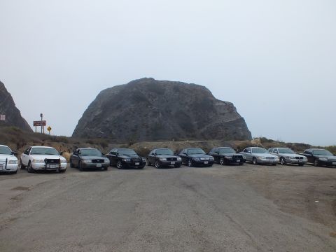 2003 Ford Crown Victoria - row of full-size sedans faces the camera in a gravel lot beneath a large rocky dome under foggy sky.