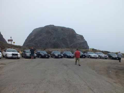 2003 Ford Crown Victoria - Two people stand among a row of front-facing full-size sedans parked in a gravel lot beneath a large rock formation.